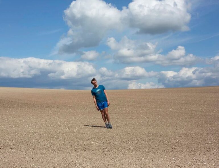 photographic image on a cloudy sunny day, there is a man standing at an angle in a big ploughed field.
